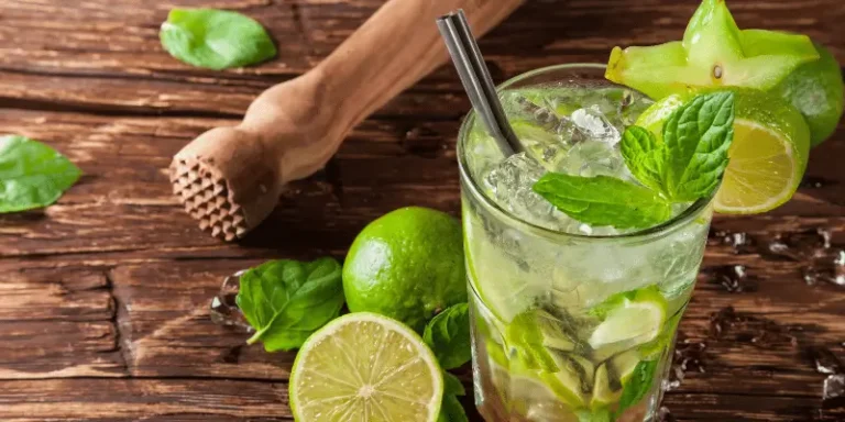 Imagem de um mojito, em uma mesa, junto com limões ao redor