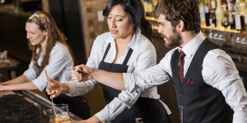 Imagem mostra um barman ajudando uma bartender na criação d de um drink.