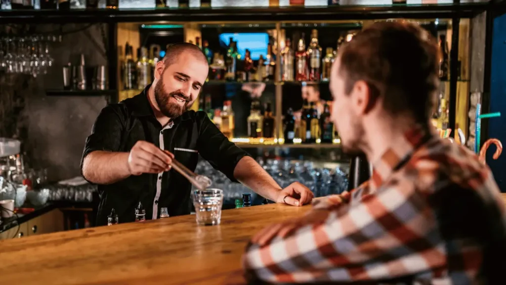 Barman preparando uma bebida, com um sorriso na cara e cliente olhando no balcão