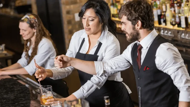 Um homem e duas mulheres realizando serviço de barman. Os três são brancos e estão preparando bebidas no balcão.