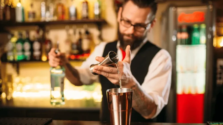 Barman faz cocktail no bar. Ele é um homem branco e usa camisa branca e um colete preto.