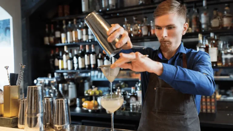 Barman usa cocktail shaker para fazer um dtink. Ele é de pele branca, loiro e está num bar.