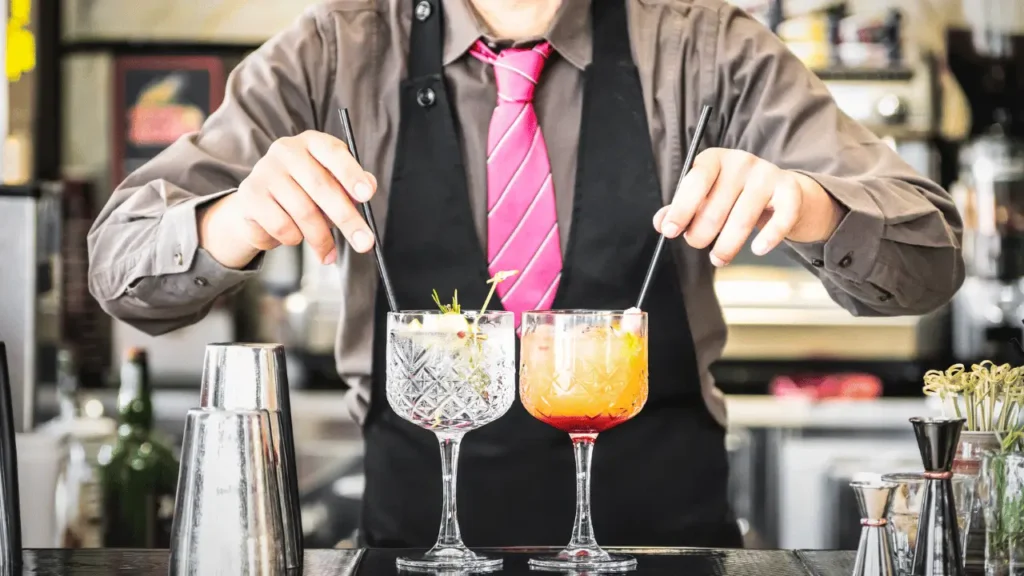 Barman colocando canudo em dois drinks. Ele está vestindo uma camisa social cinza, gravata rosa e avental preto. 