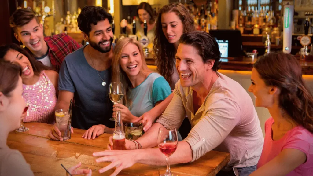 Pessoas na mesma mesa de um bar, com bebidas na mesa. São dois homens e duas mulheres. Em segundo plano, uma bartender no balcão.