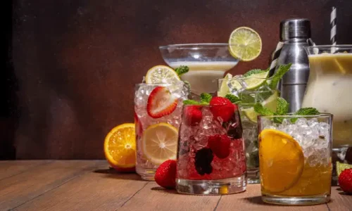 Imagem mostra muitos ingredientes, com sumos e frutas, para um mocktail, em uma mesa.